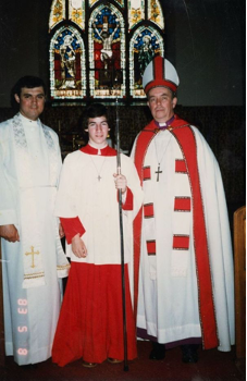 Fr Young in white robes beside a server and bishop.